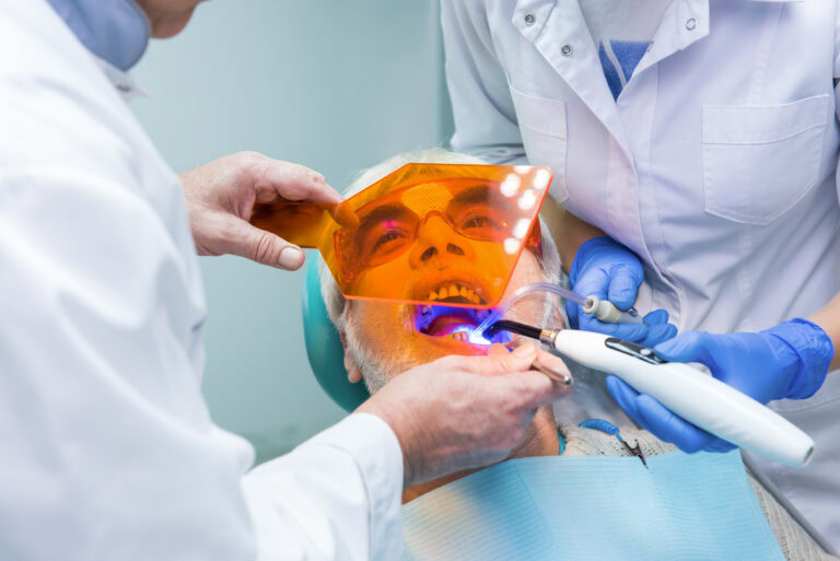 Dentist performing gum disease treatment on an older male patient using UV light and protective eyewear